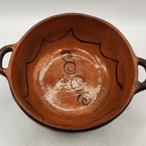 May include: A terracotta serving bowl with two handles. The bowl has a brown glaze and a black design on the inside. The design includes a sunburst pattern and the word "Tacos" written in cursive.
