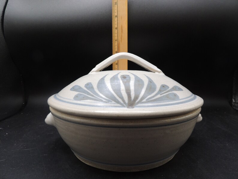 May include: A white ceramic casserole dish with a lid. The lid features a blue and white floral design and a white handle. The dish has two small handles on the sides.