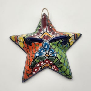 May include: A colorful, star-shaped ceramic ornament with a detailed floral design. The ornament features a vibrant mix of orange, green, yellow, and blue hues, with a dark blue border and a natural fiber loop for hanging. The star is approximately 6 inches across.