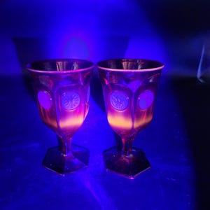 May include: Two amber-colored glass goblets with hexagonal bases and embossed circular medallions on the bowls. The glass glows with a bright orange hue under ultraviolet light, highlighting the intricate sun and figure designs on the medallions.