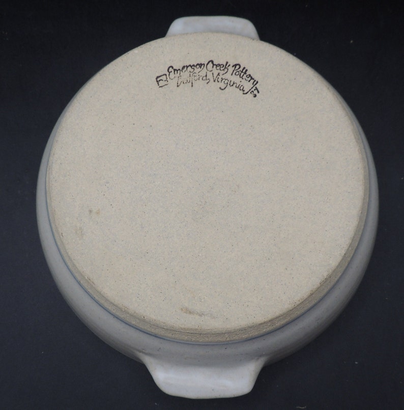 May include: A white ceramic baking dish with two handles. The bottom of the dish has a blue rim and the words "Emerson Creek Pottery, Bluford, Virginia" in black.