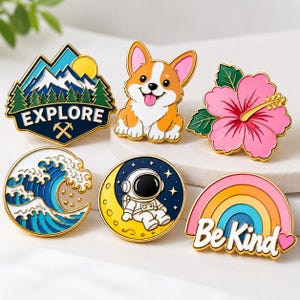 Può includere: Una collezione di sei spille smaltate colorate raffiguranti un paesaggio montano con il testo EXPLORE, un corgi, un fiore di ibisco rosa, un'onda oceanica blu, un astronauta su una luna crescente gialla e un arcobaleno con il testo Be Kind.