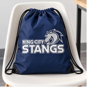May include: A navy blue drawstring backpack featuring a white and silver graphic of a horse head and the text KING CITY STANGS. The bag has black cord straps and black reinforced corners with metal grommets for the drawstring closure mechanism.