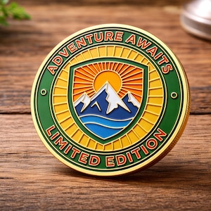 Puede incluir: Una moneda redonda de color dorado con un borde verde y amarillo. La moneda presenta un diseño de montaña y sol dentro de un escudo, con las palabras "ADVENTURE AWAITS" y "LIMITED EDITION" en rojo. La moneda está sobre una superficie de madera.
