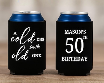 May include: Two black can coolers with white text. One says "A cold one for the old one" in cursive. The other says "Mason's 50th Birthday". The cans inside are blue.