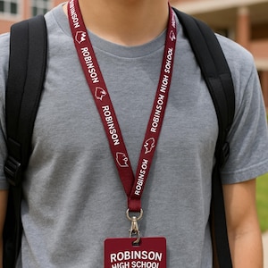 May include: A maroon lanyard with the text "ROBINSON HIGH SCHOOL" and "ROBINSON" repeated. The lanyard has a silver clasp and a maroon ID card holder. The person is wearing a gray t-shirt and a black backpack.