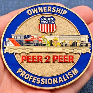 Puede incluir: Una moneda redonda de color dorado con un borde azul. La moneda presenta un tren con un diseño de bandera estadounidense, las palabras "OWNERSHIP", "PEER 2 PEER" y "PROFESSIONALISM". El logotipo de Union Pacific está en la parte superior.
