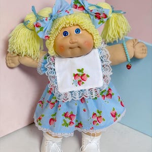 May include: A doll with light yellow yarn hair styled in pigtails with blue strawberry-patterned bows. The doll wears a light blue dress with a strawberry print, a white lace-trimmed bib, white shoes, and a light blue beaded strawberry bracelet.