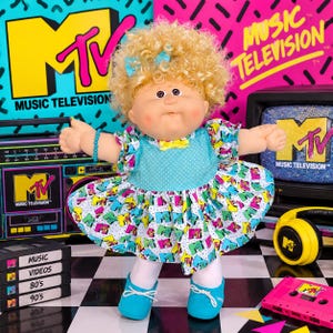 Peut inclure: Une poupée aux cheveux blonds bouclés avec un nœud bleu, portant un corsage bleu sarcelle à pois et une jupe blanche à motif logo MTV coloré. La poupée porte des leggings blancs et des chaussures bleu sarcelle, avec un bracelet bleu et un nœud jaune.