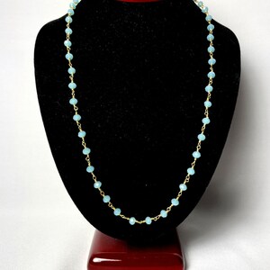 K&ouml;nnte beinhalten: Eine zarte Halskette mit kleinen, hellblauen Perlen auf einer goldfarbenen Kette. Die Halskette wird auf einer schwarzen B&uuml;ste vor wei&szlig;em Hintergrund pr&auml;sentiert. Die Perlen sind gleichm&auml;&szlig;ig verteilt.