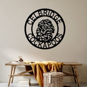 Puede incluir: Arte de pared de metal negro con un diseño de perro Cockapoo dentro de un marco circular. El texto "CELBRIDGE COCKAPOOS" está dispuesto alrededor de la imagen del perro. La obra de arte está montada en una pared de color neutro.