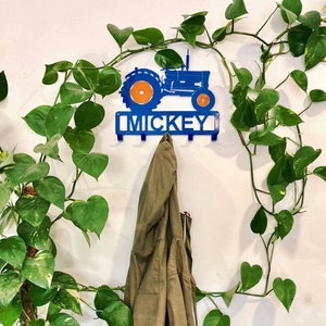 May include: Blue metal tractor wall hook with the name "MICKEY" in blue letters. The tractor has orange wheels and is mounted on a white wall. A green jacket is hanging on one of the hooks.