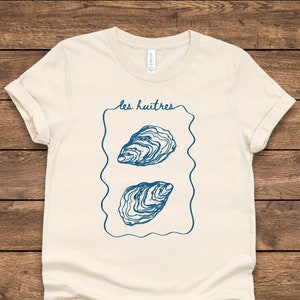 Puede incluir: Una camiseta de color crema con un gráfico azul de dos ostras y el texto "les huitres" en una fuente cursiva.