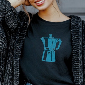 May include: Black t-shirt with a turquoise illustration of a coffee maker. The graphic is centered on the chest. The person is wearing blue jeans and a dark grey cardigan.