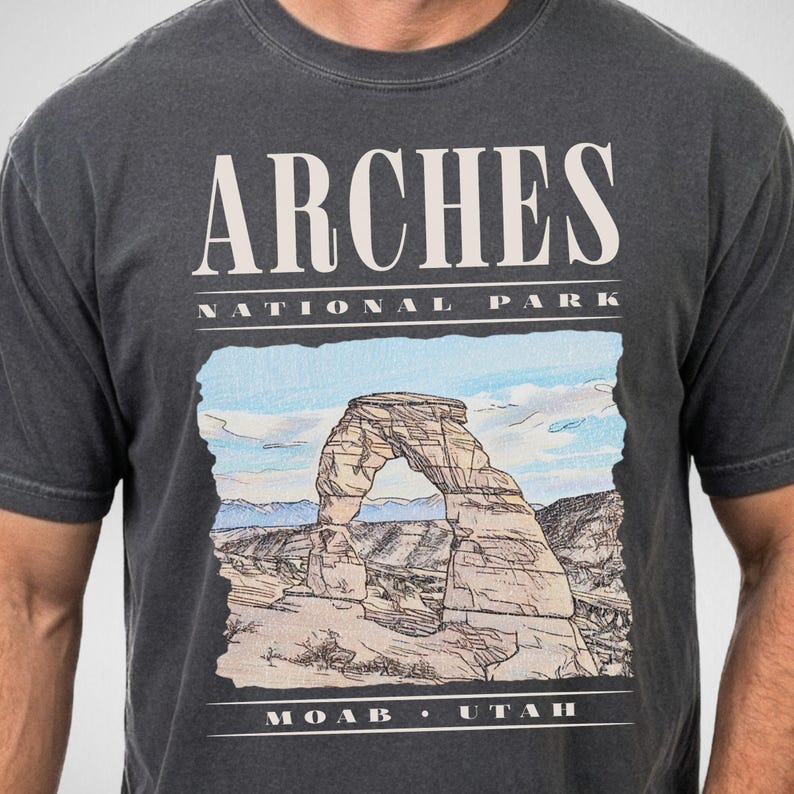 A person wears a Comfort Colors t-shirt in the color Pepper with a vintage graphic of Delicate Arch and the text, "Arches National Park, Moab Utah."