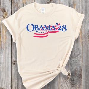 Puede incluir: Una camiseta beige con un gráfico azul y rojo que dice "OBAMA '28" con estrellas y rayas rojas y blancas.