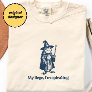 以下が含まれることがあります： クリーム色のTシャツ。ネイビーブルーのイラストで、魔法使いの格好をしたカエルが杖を持っています。「My liege, I'm spiraling」の文字が下にあります。丸首で半袖のシャツです。