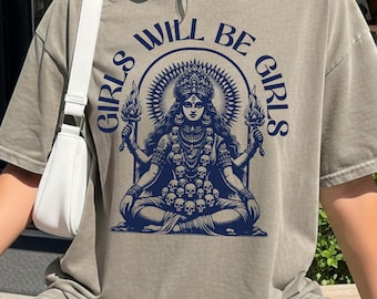 May include: A beige t-shirt featuring a dark blue graphic print of a multi-armed deity figure seated in a meditative pose, adorned with a skull necklace and holding torches, surrounded by the text "GIRLS WILL BE GIRLS" in an arch.