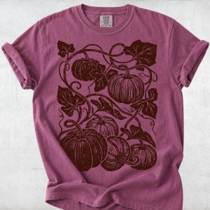 May include: A mauve short-sleeve t-shirt featuring a dark brown graphic print of multiple pumpkins with curling vines and large leaves. The shirt has a crew neckline and a Comfort Colors brand label printed on the inside of the collar area.