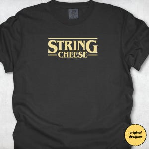 Puede incluir: Camiseta negra con el texto "STRING CHEESE" en amarillo, con una fuente de estilo retro. La camiseta tiene cuello redondo y mangas cortas. Una pequeña etiqueta es visible en la parte superior, y una pegatina de "diseñador original" en la parte inferior derecha.