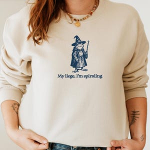 May include: Cream-colored sweatshirt featuring a navy blue illustration of a frog dressed as a wizard, holding a wand. The text "My liege, I'm spiraling" is printed below the graphic. The sweatshirt has long sleeves and a crew neck.