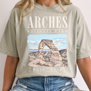 Retro Arches National Park graphic tee featuring a vintage watercolor image of Delicate Arch with bold nineties style cream typography reading "Arches National Park, Moab, Utah" printed on a Sandstone Comfort Colors tee.