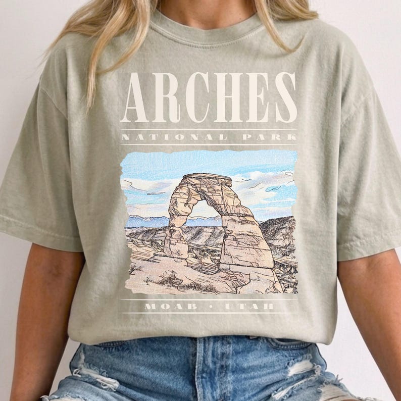 Retro Arches National Park graphic tee featuring a vintage watercolor image of Delicate Arch with bold nineties style cream typography reading "Arches National Park, Moab, Utah" printed on a Sandstone Comfort Colors tee.