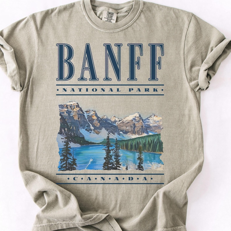 A Sandstone Comfort Colors t-shirt with dark blue vintage text reading "Banff National Park" and "CANADA," with a graphic illustration of Moraine Lake.