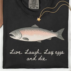 May include: A black t-shirt featuring a graphic illustration of a fish with a pinkish side and grey scales. Below the fish, the text reads, "Live. Laugh. Lay eggs and die." A gold-colored necklace with a small star charm rests on the shirt collar.
