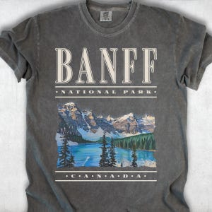 A dark grey Comfort Colors t-shirt with off-white vintage text reading "Banff National Park" and "CANADA," with a graphic illustration of Moraine Lake.