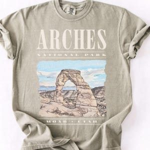 Retro Arches National Park graphic tee featuring a distressed watercolor image of Delicate Arch with bold cream typography reading "Arches National Park, Moab, Utah" printed on a Sandstone Comfort Colors tee.