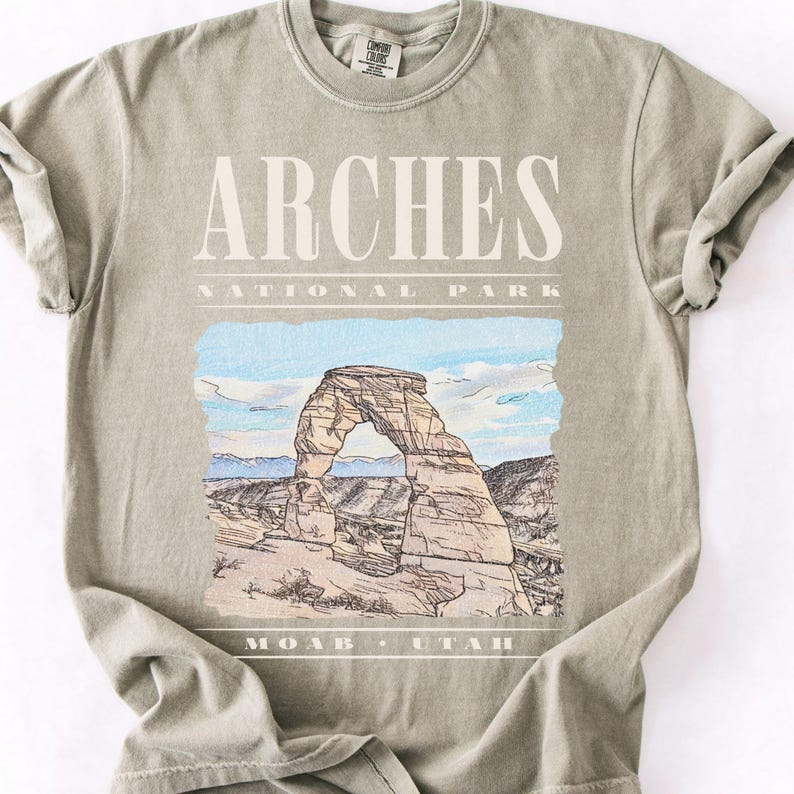 Retro Arches National Park graphic tee featuring a distressed watercolor image of Delicate Arch with bold cream typography reading "Arches National Park, Moab, Utah" printed on a Sandstone Comfort Colors tee.