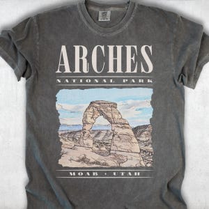 Retro Arches National Park graphic tee featuring a distressed watercolor image of Delicate Arch with bold cream typography reading "Arches National Park, Moab, Utah" printed on a Pepper Comfort Colors tee.