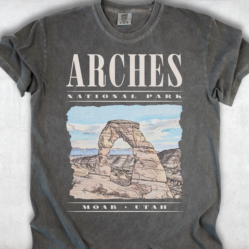 Retro Arches National Park graphic tee featuring a distressed watercolor image of Delicate Arch with bold cream typography reading "Arches National Park, Moab, Utah" printed on a Pepper Comfort Colors tee.