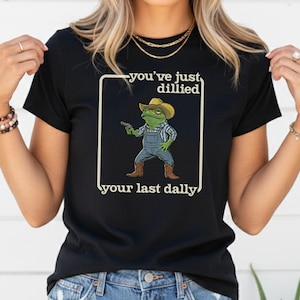 Puede incluir: Camiseta negra con una rana de dibujos animados con sombrero de vaquero, peto y botas, sosteniendo una pistola. El texto dice "you've just dillied your last dally."