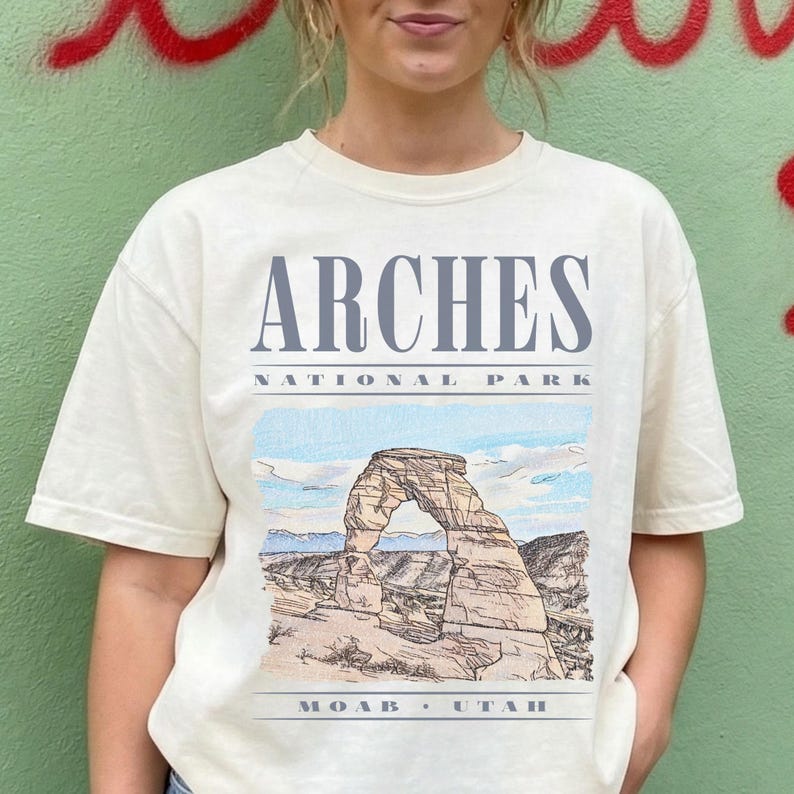 Retro Arches National Park graphic tee featuring a distressed watercolor image of Delicate Arch with bold blue-grey typography reading "Arches National Park, Moab, Utah" printed on an Ivory Comfort Colors tee.
