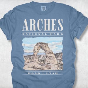 Retro Arches National Park graphic tee featuring a distressed watercolor image of Delicate Arch with bold cream typography reading "Arches National Park, Moab, Utah" printed on a Blue Jean Comfort Colors tee.
