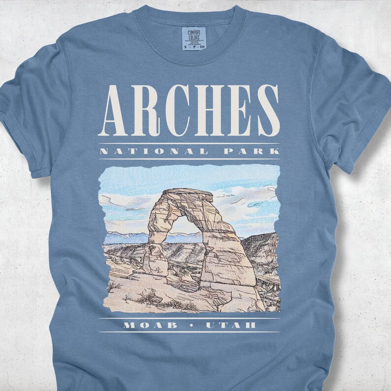 Retro Arches National Park graphic tee featuring a distressed watercolor image of Delicate Arch with bold cream typography reading "Arches National Park, Moab, Utah" printed on a Blue Jean Comfort Colors tee.