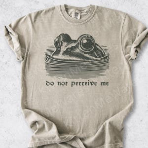 May include: A beige short-sleeve t-shirt featuring a dark gray graphic of a frog partially submerged in water. Below the frog, the text reads "do not perceive me" in a lowercase serif font. The shirt has a crew neckline and a standard fit.