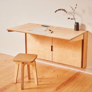 Peut inclure: Un bureau mural en bois clair avec une surface rabattable et deux portes de placard, accompagné d'un tabouret en bois assorti à quatre pieds. Le bureau porte un smartphone, deux stylos, deux papiers et un vase sombre avec une branche séchée.