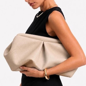 May include: A beige clutch bag with a soft, gathered, pleated texture and a structured frame opening. The bag features a pebbled surface finish and is held horizontally. A gold-toned bangle bracelet is visible on the wrist holding the clutch.