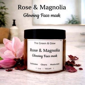 May include: A jar of Rose & Magnolia Glowing Face mask with a black lid and a white label. The label includes the text "The Green & Glow" and product details. Pink magnolia flowers and petals are arranged around the jar. The jar contains 5 oz of product.