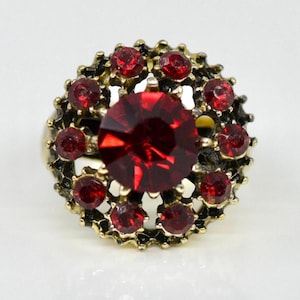 May include: A vintage-style ring with a large, faceted, deep red gemstone at the center, surrounded by smaller red stones. The ring has a gold-toned metal setting with an ornate, floral-like design. The ring is set against a white background.