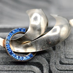 May include: A vintage silver-tone brooch with a swirling design. The brooch features a circular element encrusted with square, bright blue gemstones. The design includes two curved, wing-like shapes. A classic piece of jewelry.