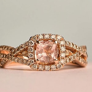 Puede incluir: Anillo de oro rosa con una piedra preciosa rosa pálido cuadrada, rodeada de pequeñas piedras transparentes. La banda tiene un diseño de vástago dividido con más piedras pequeñas. El anillo está sobre un fondo suave y neutro.