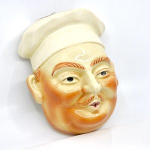May include: A vintage ceramic chef head wall hanging. The chef has a cream-colored toque and face with orange and brown accents. The chef has a mustache and is looking slightly to the side.