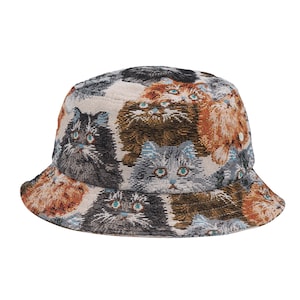 May include: A bucket hat featuring a repeating pattern of cat faces in shades of black, brown, gray, and orange. The hat has a wide brim and is made of a woven fabric. The cat faces have blue eyes.