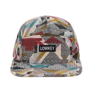 May include: A five-panel cap with a geometric pattern in shades of beige, red, yellow, and teal. The cap features a flat brim and a small black rectangular patch with the word "LOWKEY" in white.