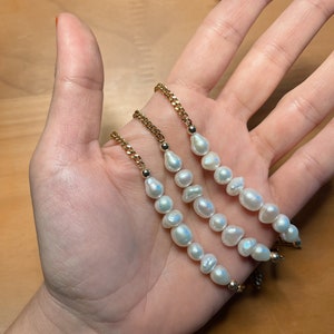 Puede incluir: Tres pulseras de cadena dorada, cada una adornada con perlas de agua dulce blancas irregulares. Las pulseras se muestran en una mano, mostrando el dise&ntilde;o delicado y el brillo de las perlas. Las cadenas tienen una peque&ntilde;a cuenta dorada en cada extremo.