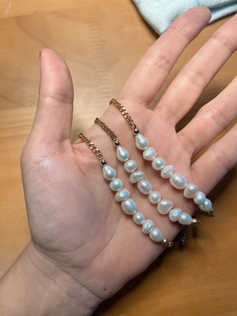 Puede incluir: Tres pulseras de cadena dorada, cada una adornada con perlas de agua dulce blancas irregulares. Las pulseras se muestran en una mano, mostrando el dise&ntilde;o delicado y el brillo de las perlas. Las cadenas tienen una peque&ntilde;a cuenta dorada en cada extremo.